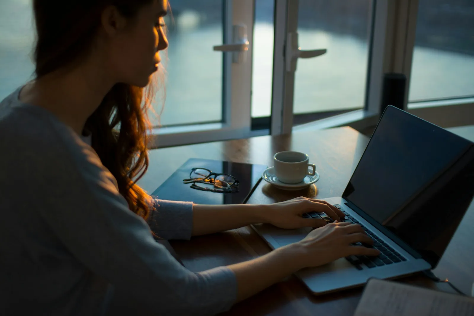 A person at a desk by a window, hands on a laptop, a coffee beside them